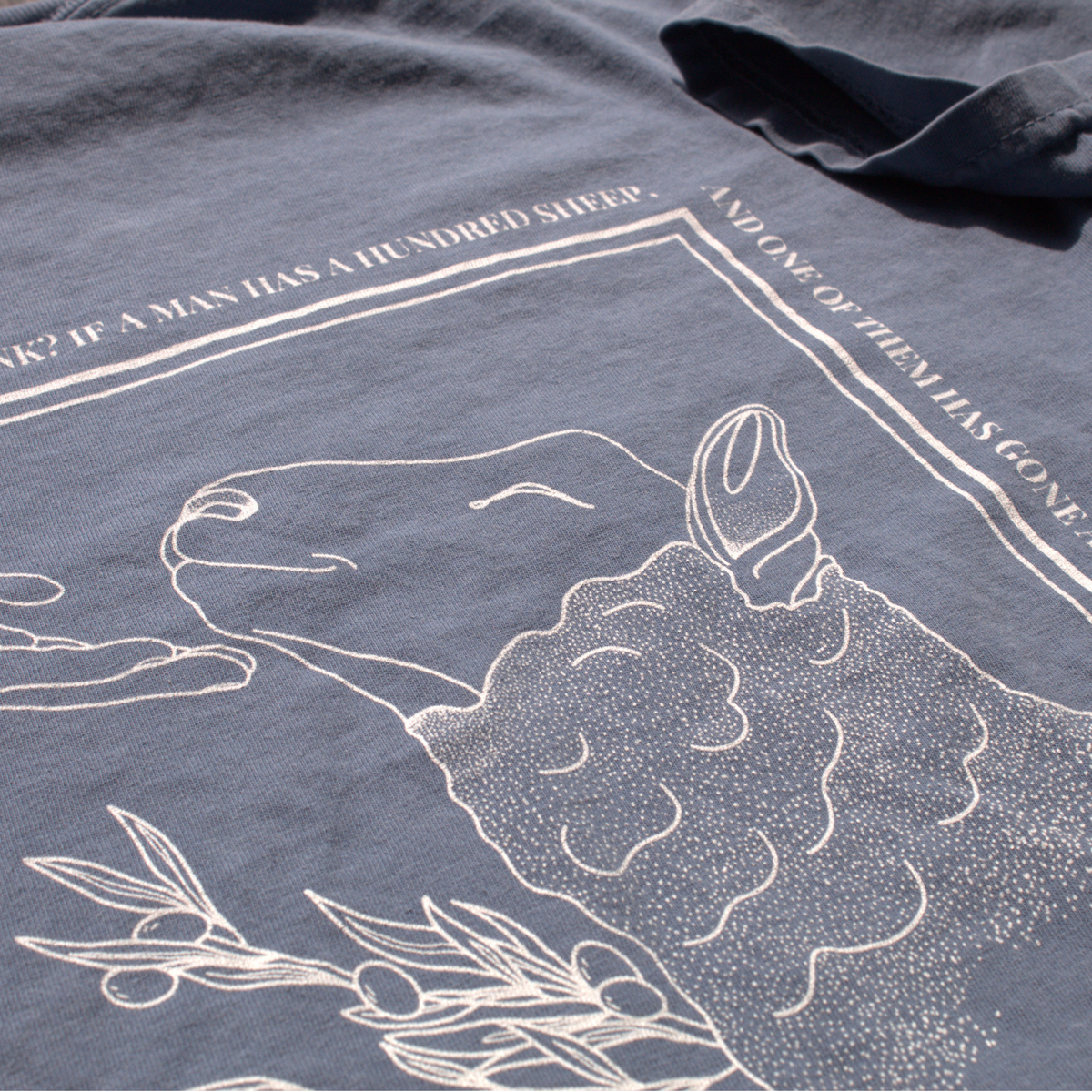 close up of the Dusty blue shirt with white line art of a sheep being comforted by the hand of Jesus.