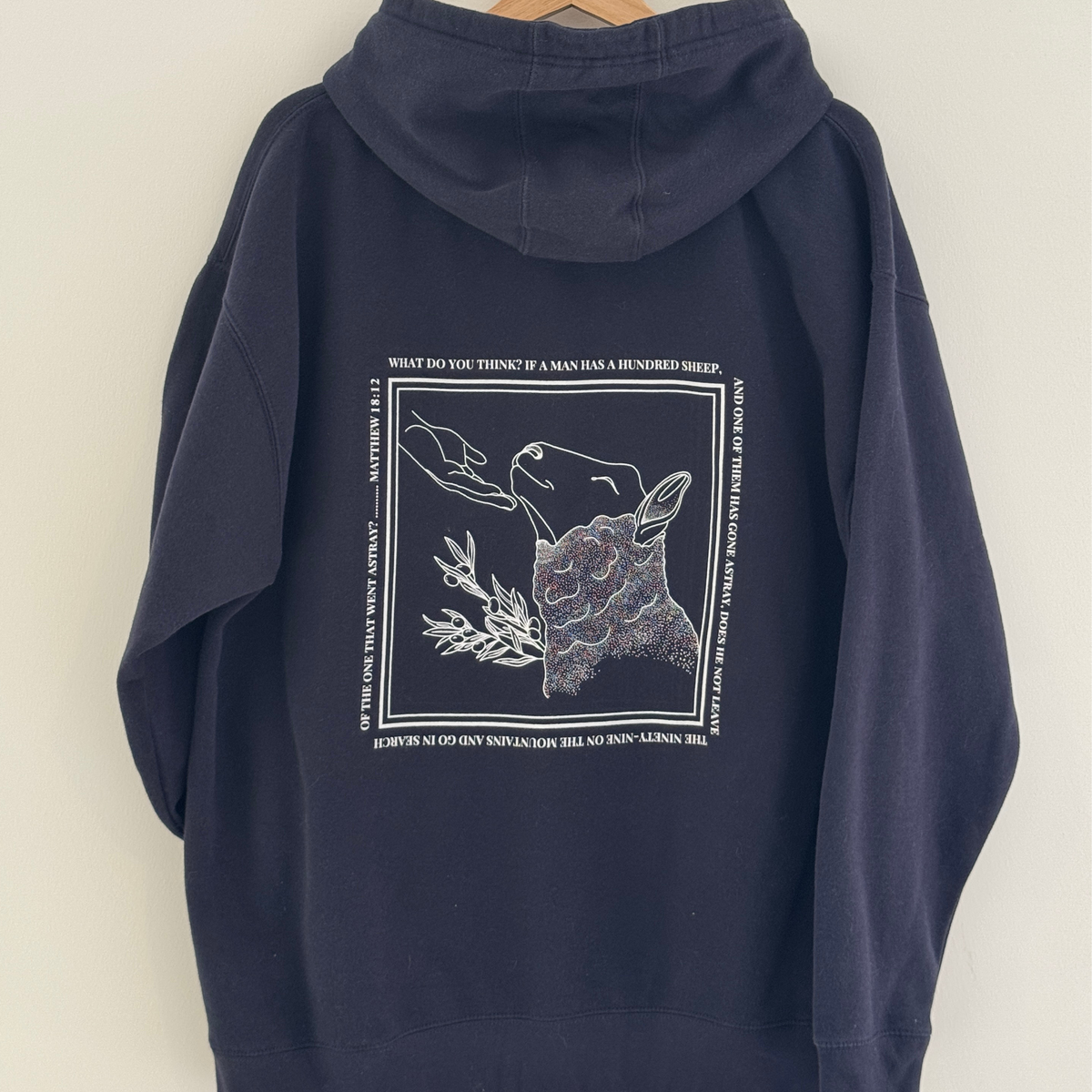 Navy blue hoodie with a graphic design on the back depicting a sheep being comforted by the hand of Jesus with the verse Matthew 18:12 surrounding it 
