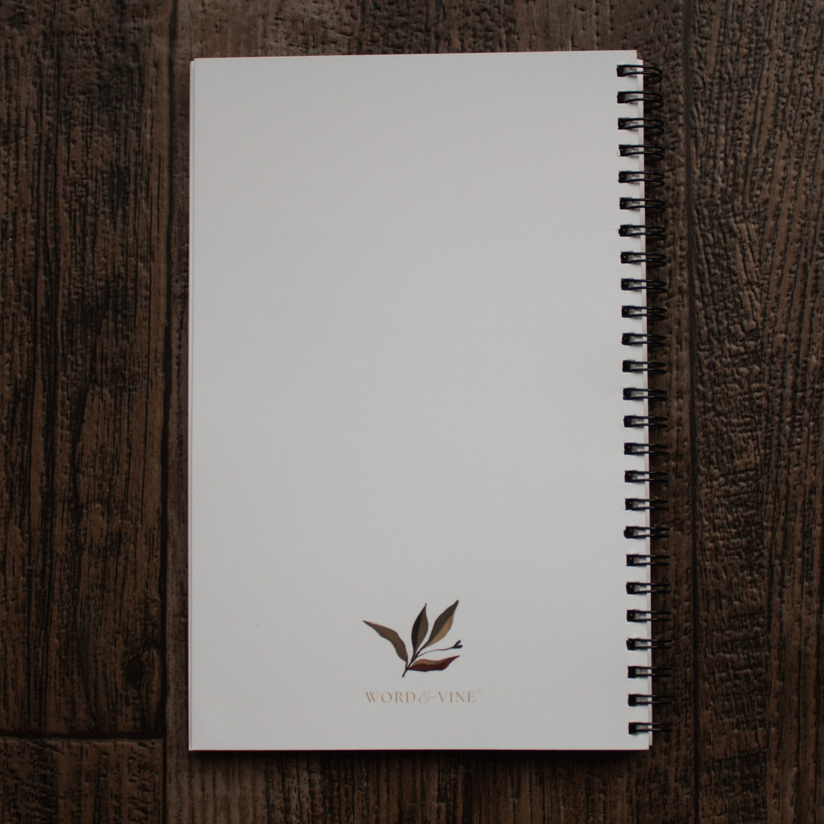 A white spiral-bound notebook back cover with a small leaf illustration and the text 'WORD and VINE' at the bottom, placed on a dark wood background.