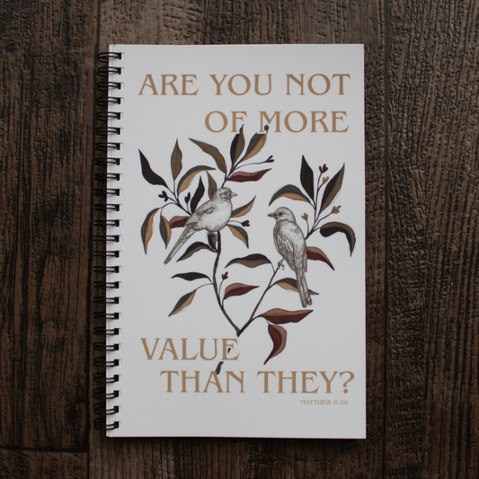 A spiral-bound notebook with a white cover lies flat on a dark wood surface. The cover features a botanical illustration of two small birds perched on branches with leaves, accompanied by the text 'ARE YOU NOT OF MORE VALUE THAN THEY?' and a small Bible verse at the bottom right. Matthew 6:26