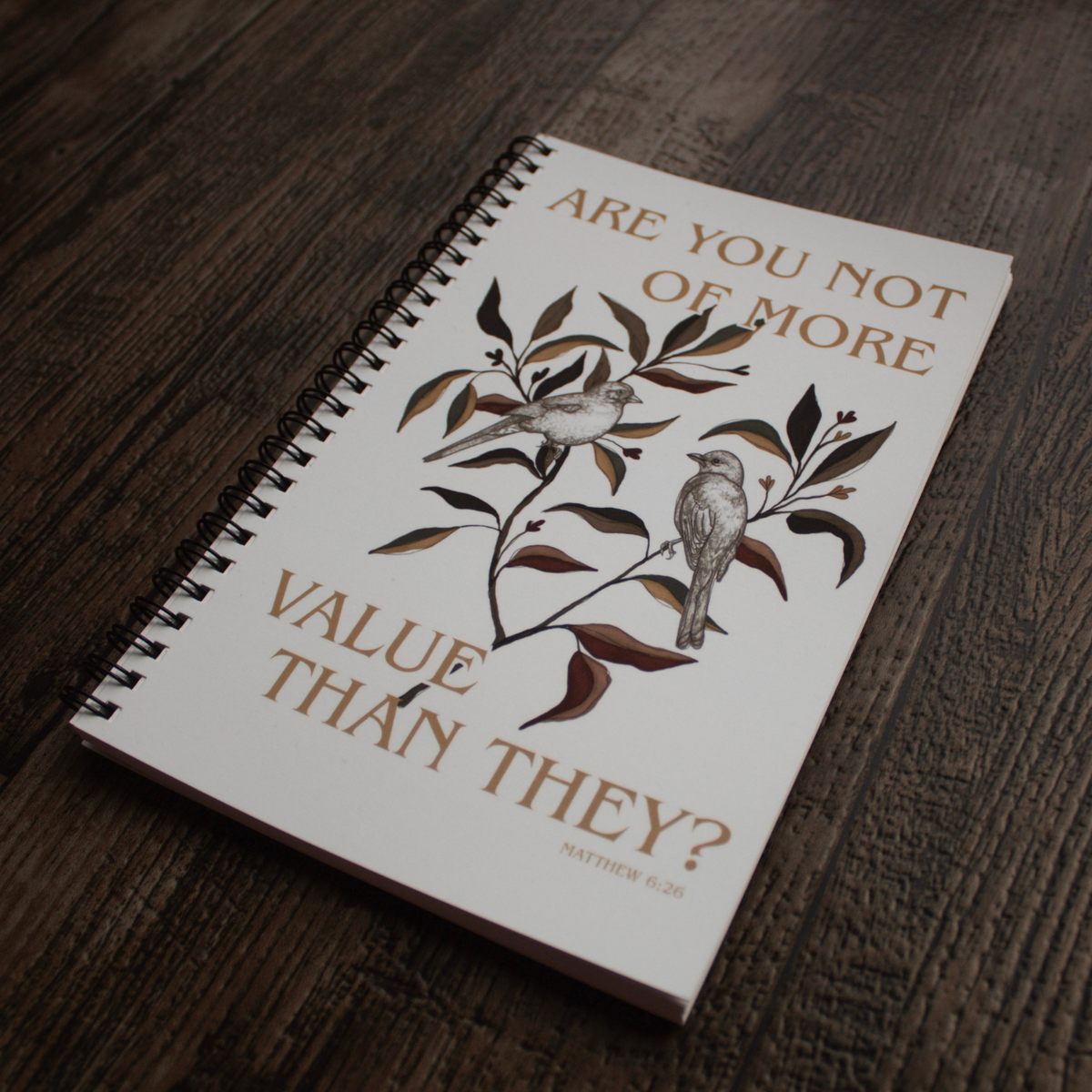 Image taken at another angle: A spiral-bound notebook with a white cover lies on a dark wood surface. The cover features a brown and gray illustration of two birds on a branch with the text 'ARE YOU NOT OF MORE VALUE THAN THEY?' and 'MATTHEW 6:26'.