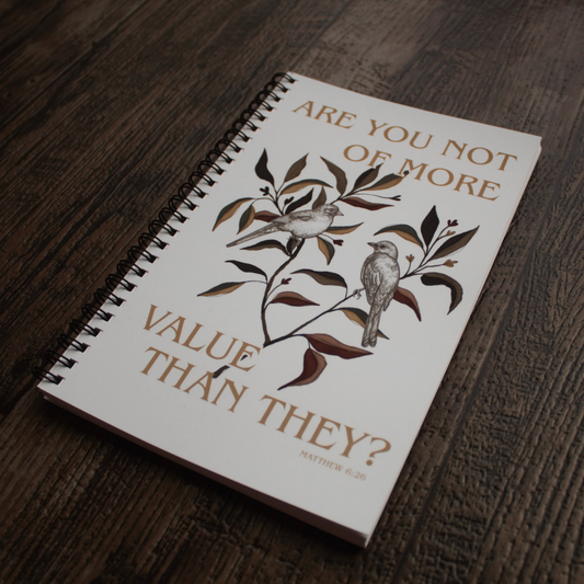 Image taken at another angle: A spiral-bound notebook with a white cover lies on a dark wood surface. The cover features a brown and gray illustration of two birds on a branch with the text 'ARE YOU NOT OF MORE VALUE THAN THEY?' and 'MATTHEW 6:26'.