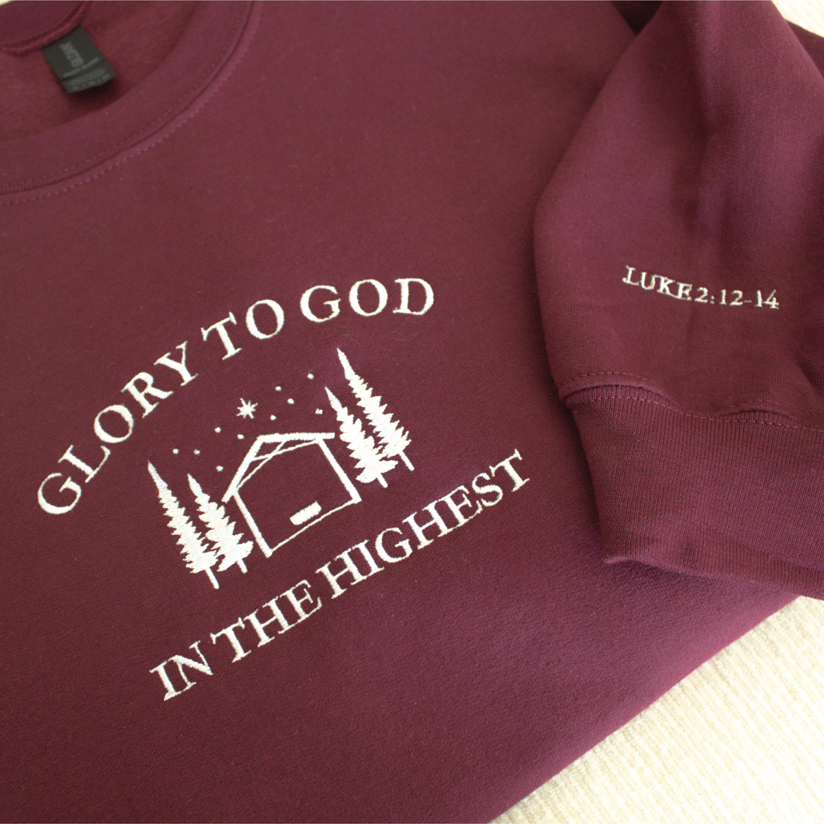 Maroon sweatshirt with embroidered 'Glory to God in the Highest' text and simple manger and stable in between trees with stars and the star of David above. Sweatshirt sleeve features 'Luke 2:12-14' white embroidered text 
