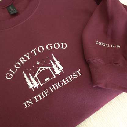 Maroon sweatshirt with embroidered 'Glory to God in the Highest' text and simple manger and stable in between trees with stars and the star of David above. Sweatshirt sleeve features 'Luke 2:12-14' white embroidered text 