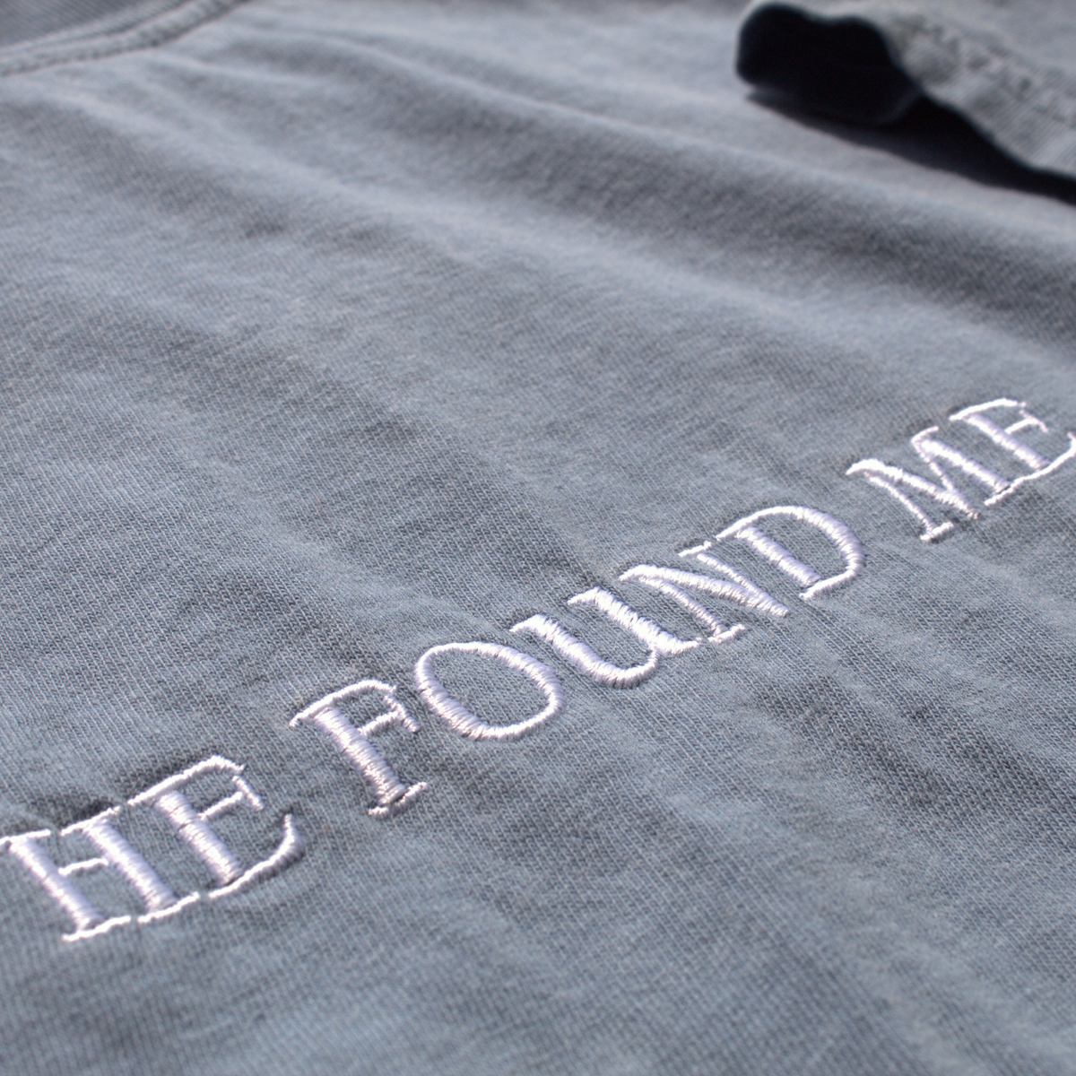 close up of the Dusty blue tee with 'HE FOUND ME' embroidered in white thread