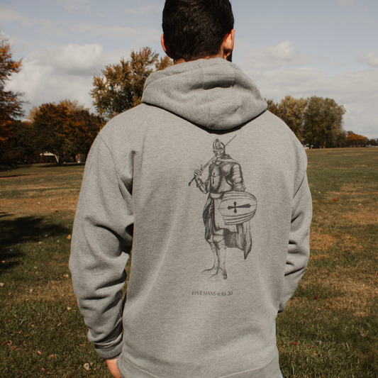 Person wearing a gray hoodie with a armored knight design on the back with Ephesians 6:10-20 written at the bottom, standing in an outdoor setting.