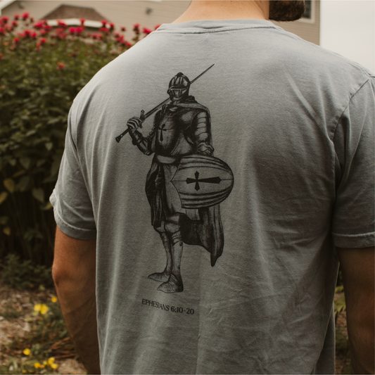 Person wearing a gray t-shirt with a black graphic design of an armored knight on the back with the text 'Ephesians 6:10-12' , standing outdoors.
