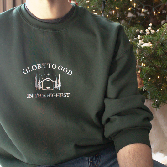 Forest Green sweatshirt with 'Glory to God in the Highest' text and Embroidered 'Glory to God in the Highest' text and simple manger and stable in between trees with stars and the star of David above. Worn by a person in front of a decorated Christmas tree.