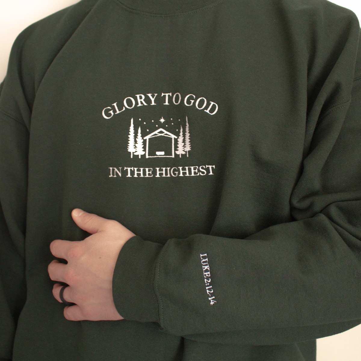 Forest Green modeled sweatshirt featuring embroidered 'Glory to God in the Highest' text and simple manger and stable in between trees with stars and the star of David above. Sweatshirt sleeve features 'Luke 2:12-14' white embroidered text 