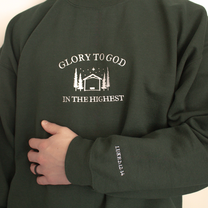 Forest Green modeled sweatshirt featuring embroidered 'Glory to God in the Highest' text and simple manger and stable in between trees with stars and the star of David above. Sweatshirt sleeve features 'Luke 2:12-14' white embroidered text 