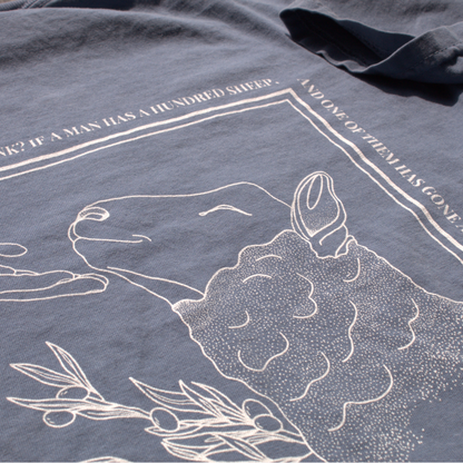 close up of the Dusty blue shirt with white line art of a sheep being comforted by the hand of Jesus. 