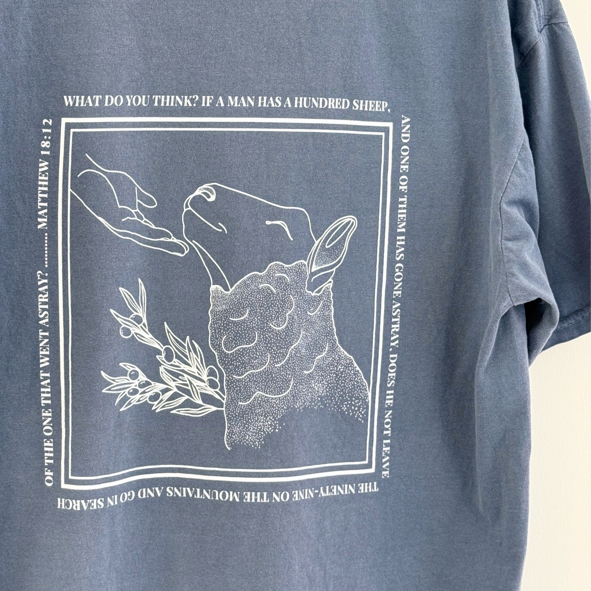 Dusty Blue t-shirt with white graphic of a sheep being comforted by the hand of Jesus and the verse of Matthew 18:12 surrounding it in a square