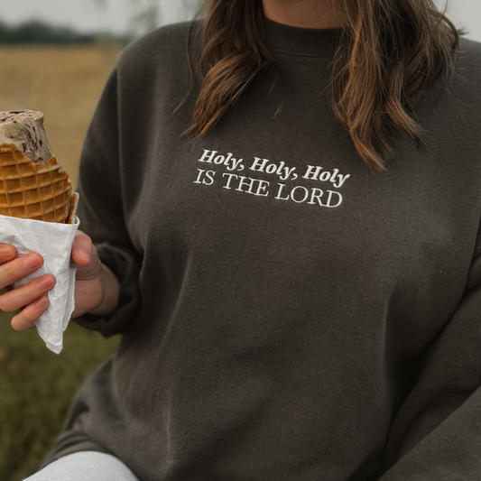 Person wearing a dark gray sweatshirt with embroidered 'Holy, Holy, Holy IS THE LORD' text, holding a waffle cone Ice cream in a field.