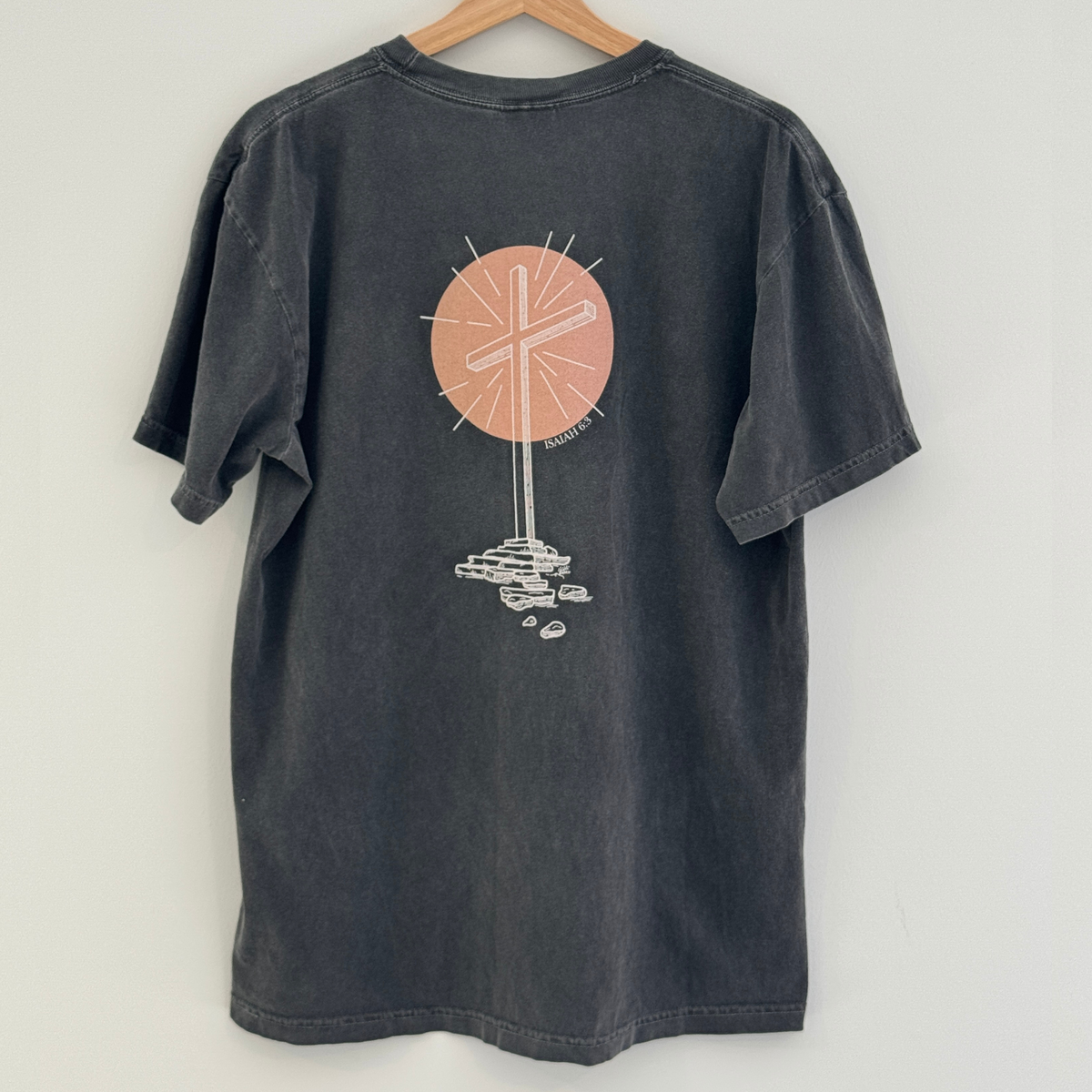 Gray t-shirt with a graphic design of an empty cross in front of a peach colored circle on the back, hanging against a white background