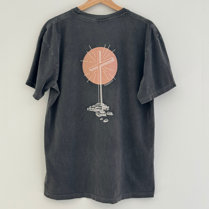 Gray t-shirt with a graphic design of an empty cross in front of a peach colored circle on the back, hanging against a white background