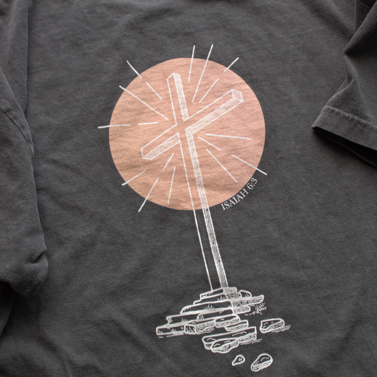 Close up of Gray t-shirt with a white graphic design of a cross and peach colored circle with text 'Isaiah 6:3' in the corner