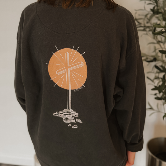 Person wearing a dark gray sweatshirt with a graphic white line art design on the back depicting the empty cross and a peach colored circle behind it