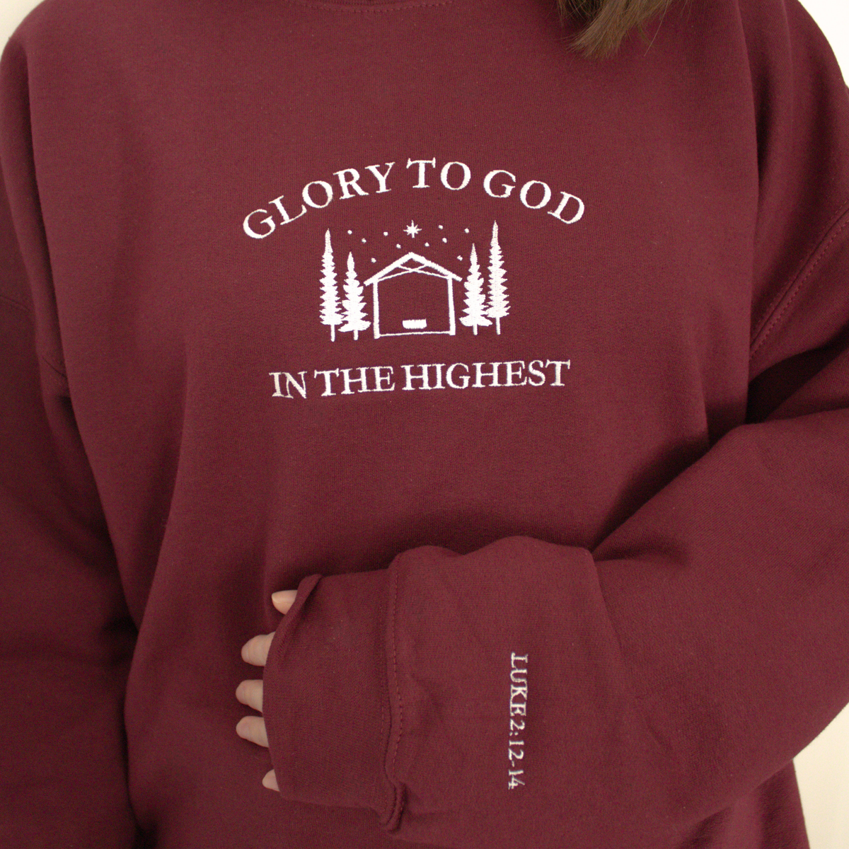 Maroon modeled sweatshirt featuring embroidered 'Glory to God in the Highest' text and simple manger and stable in between trees with stars and the star of David above. Sweatshirt sleeve features 'Luke 2:12-14' white embroidered text 