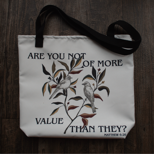 Product Image taken strait on: A white canvas tote bag with black handles, featuring a black and brown graphic of two birds on a branch with the text 'ARE YOU NOT OF MORE VALUE THAN THEY? MATTHEW 6:26', laid flat on a dark wood background.