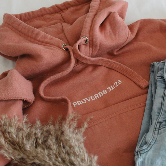 Flat Lay of Dusty Rose colored hoodie with 'Proverbs 31:25' text on a light background with blue jeans and dried plants. 