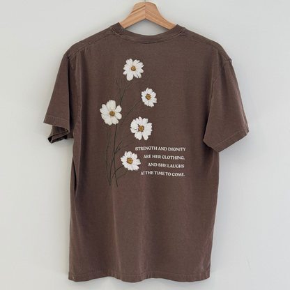 Back of Brown t-shirt with white floral design and text 'strength and dignity are her clothing, and she laughs at the time to come.' on a hanger against a white background