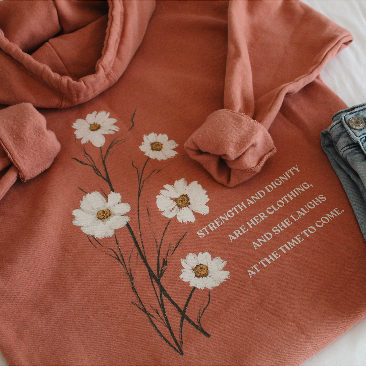 Flatlay of the back of the dusty rose colored hoodie with white floral design and text 'Strength and dignity are her clothing, and she laughs at the time to come.' on a white background with blue jeans