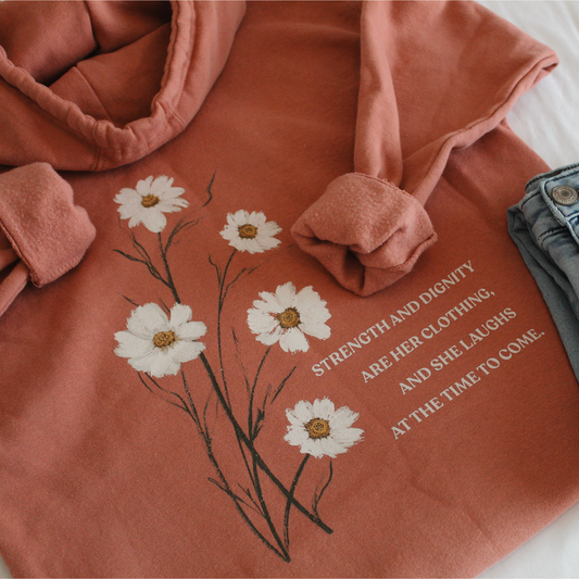 Flatlay of the back of the dusty rose colored hoodie with white floral design and text 'Strength and dignity are her clothing, and she laughs at the time to come.' on a white background with blue jeans