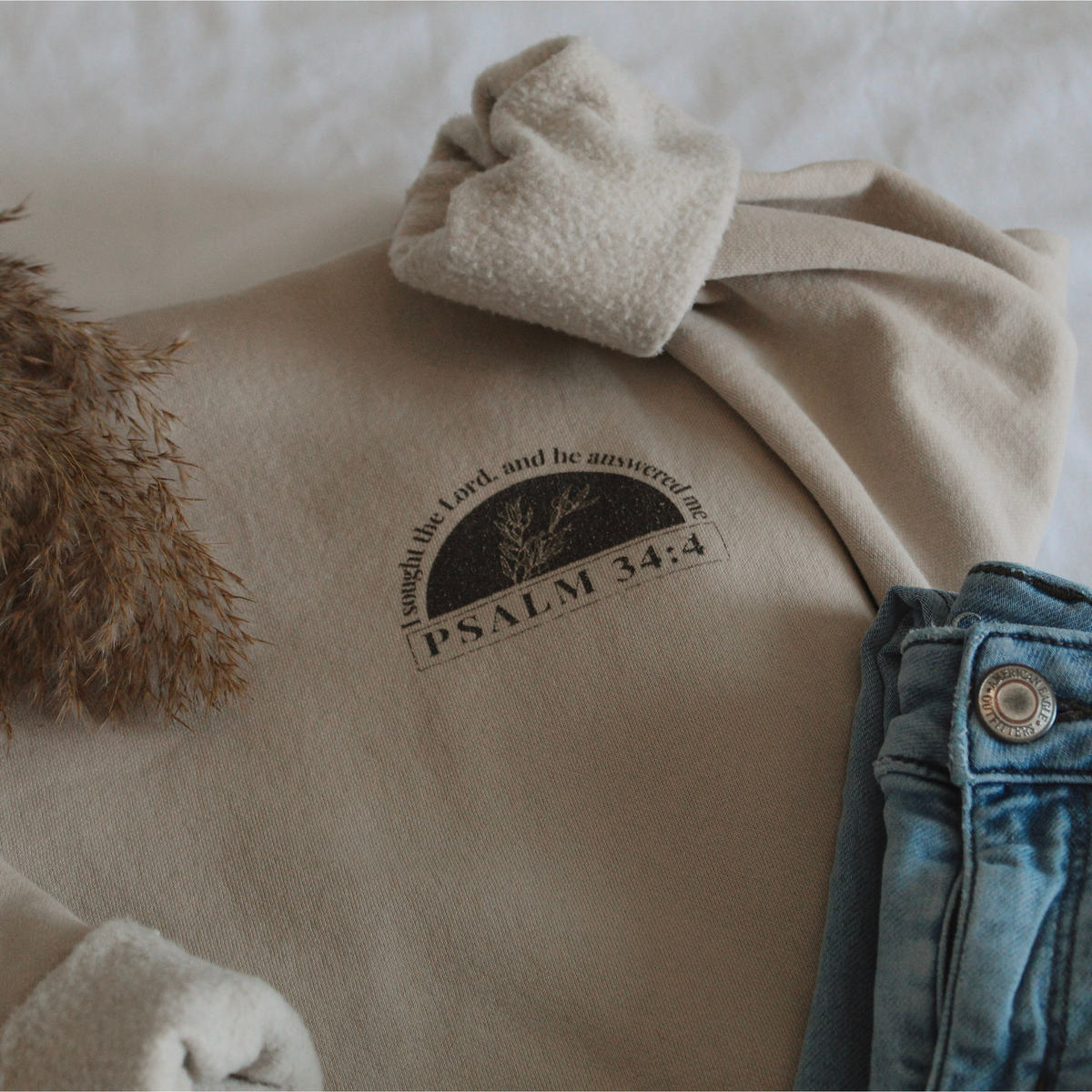 Light tan sweatshirt with a 'Psalm 34:4' text and design, paired with denim jeans and decorative grass on a neutral background.