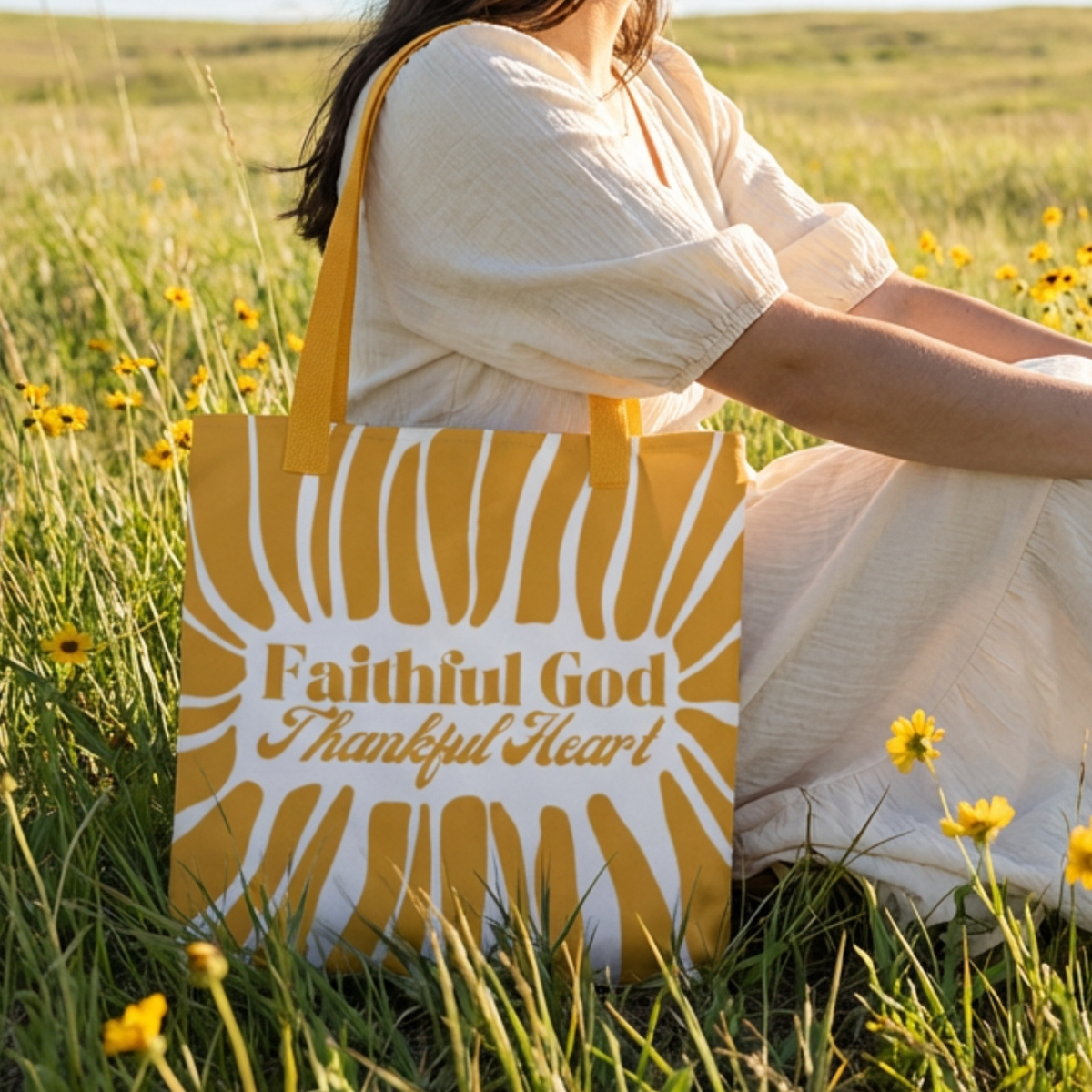 Person sitting in a field with a yellow tote bag featuring 'Faithful God Thankful Heart' text.