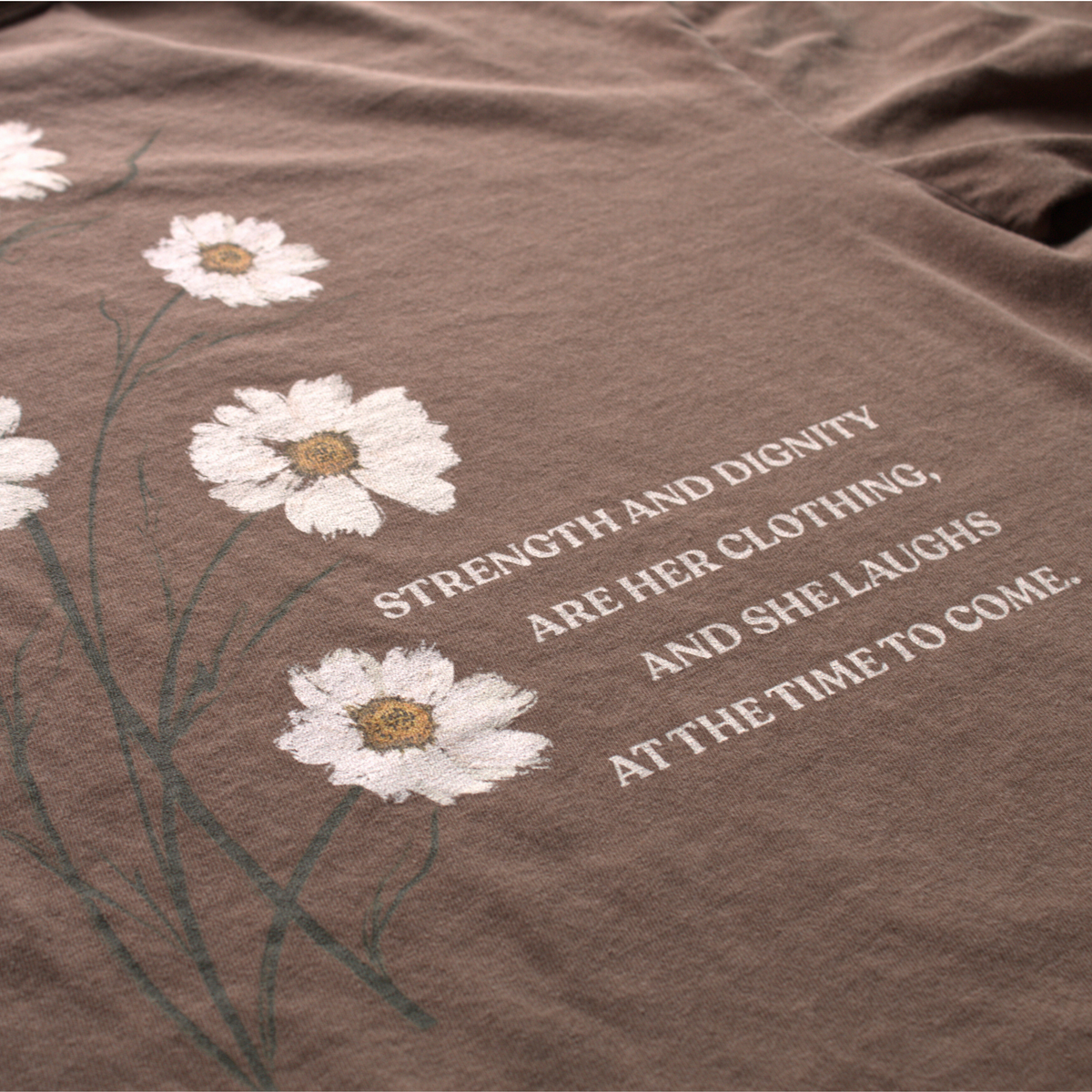 A close up of the back of the Brown t-shirt with white flowers and text reading 'strength and dignity are her clothing, and she laughs at the time to come. 