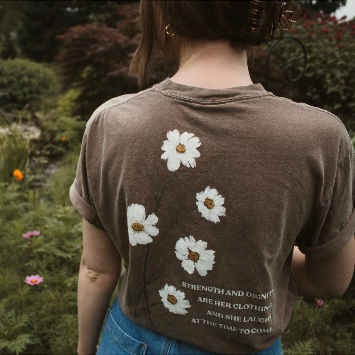 Person wearing a brown t-shirt with white floral designs and text reading 'strength and dignity are her clothing, and she laughs at the time to come.' in a garden setting