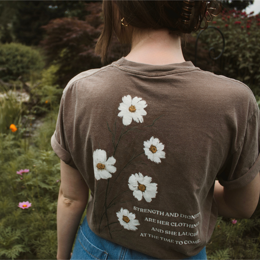 Person wearing a brown t-shirt with white floral designs and text reading 'strength and dignity are her clothing, and she laughs at the time to come.' in a garden setting